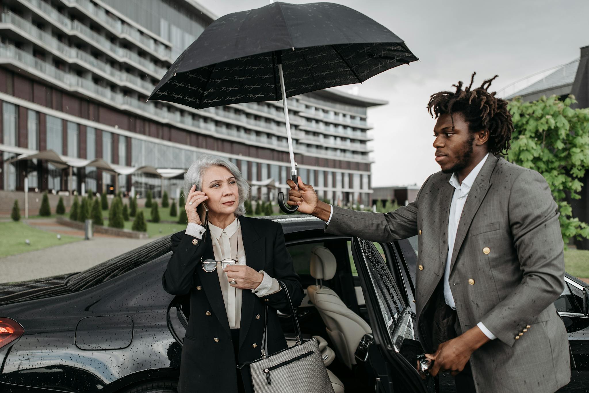 Two professionals, one holding an umbrella for the other, near a building on a rainy day.